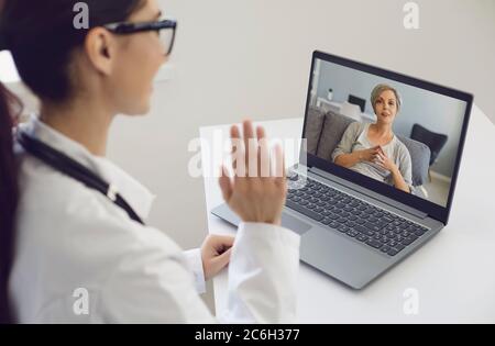 Girl doctor waves his hand consults an elderly patient woman using laptop sitting at a table in the office of the clinic. Stock Photo