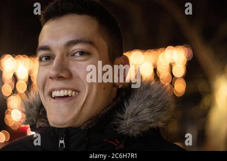 Headshot portrait of young man in winter, smiling and laughing at night with orange bokeh lights in the background Stock Photo
