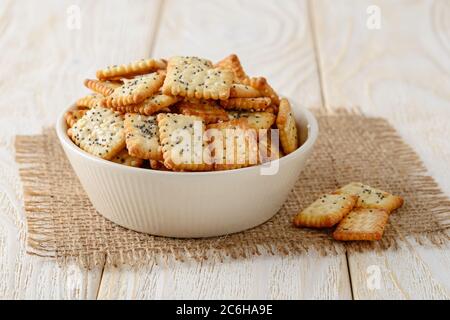 Small salty rectangular crackers with poppy and sesame seeds in a beige ceramic bowl on a white wood table. Crispy wheat flour snack and beer appetize Stock Photo