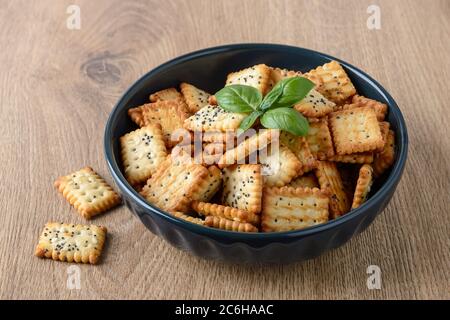 Small salty rectangular crackers with poppy and sesame seeds in a dark ceramic bowl on a brown table. Crispy wheat flour snack and beer appetizer. Stock Photo