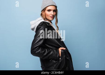 Beautiful girl with wavy hair in gray hat and black leather jacket dreamily looking in camera over blue background Stock Photo