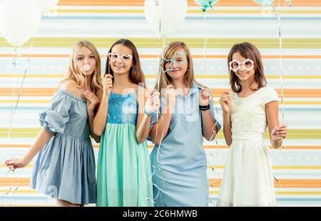 Pretty smiling teenage girls in dresses standing together with ballons at birthday party on the striped colorful background Stock Photo