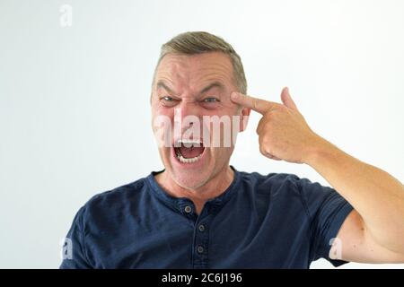 Angry man making a threatening gun gesture with his hand pointing at his own forehead while yelling at the camera in a head and shoulders portrait on Stock Photo