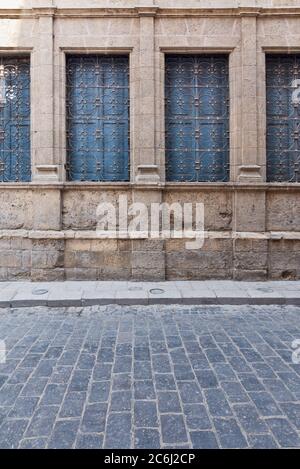 Windows with ferforge wrought iron grill and closed blue wooden shutters on old stone bricks wall, and basalt tiles road Stock Photo