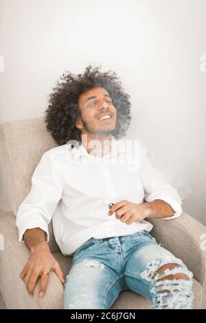 Funny young man sitting and vaping electronic cigarette at home armchair. Young smoker man resting after long working day. Alternative smoking concept Stock Photo