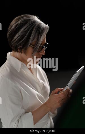 Side view of mature woman using digital tablet standing on black background. Gray haired caucasian lady Scrolls page of site or reading book in Stock Photo
