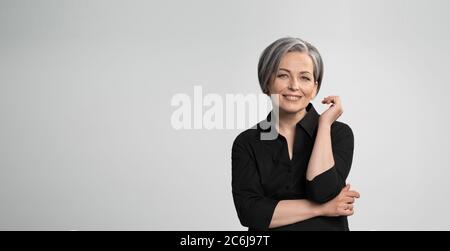 Mature Caucasian woman smiling charmingly with hand near face. Graying elegant woman with short bob hairstyle looks at camera posing on white Stock Photo