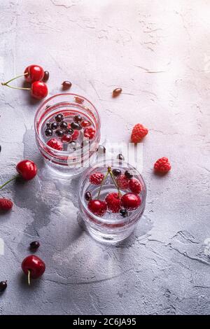 Fresh cold sparkling water drink with cherry, raspberry and currant berries in two transparent glass on stone concrete background, summer diet beverag Stock Photo