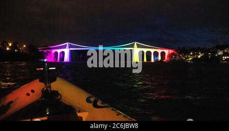 Bangor, Gwynedd, N Wales. Menai suspension Bridge over the Menai Straits is illuminated in rainbow colours to celebrate 72 years of the NHS Stock Photo