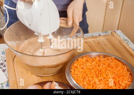 A woman is preparing a carrot cake. The woman's hand holds the mixer and mixes the ingredients. Grated carrots lie on a bowl. Egg shells lie on a plat Stock Photo