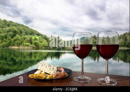 Two glasses of wine with charcuterie assortment on calm lake background in Croatia. Glass of red wine with different snacks - plate with ham, sliced Stock Photo