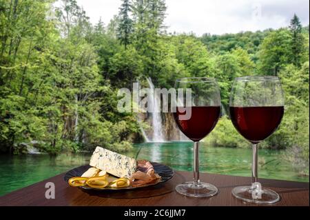 Two glasses of wine with charcuterie assortment waterfall background in Croatia. Glass of red wine with different snacks - plate with ham, sliced Stock Photo