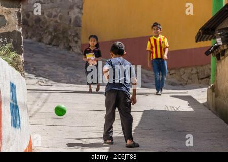 Guatemala, Solola Department, Santa Cruz la Laguna, Two young boys play soccer or football in the street as a girl in traditional dress walks by. Stock Photo