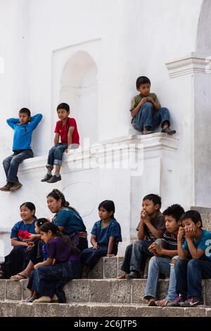 Guatemala, Solola Department, San Antonio Palopo, A Cakchiquel Mayan woman and children watch a program from the steps of the church. The woman and girls wear the traditional dress of San Antonio Palopo Guatemala, including the elaborate cinta or hair wrap, woven blue huipil blouse, faja or belt, and corte skirt.  The boys all wear modern clothing. Stock Photo