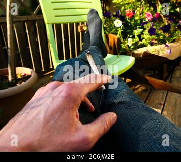 Cigarette rolled in one hand between the fingers. A person who relaxes with their legs lying on a garden chair. Concept of tobacco and smoking drugs Stock Photo