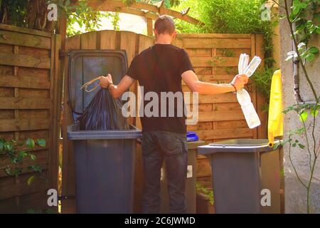 Person performing a selective sorting of household waste in recycling bins. Man putting plastic bottles in a yellow container and garbage in a bag in Stock Photo