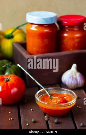 Homemade canned hot tomato sauce adjika Preserve in jars with fresh ingredients - Tomatoes, chilli pepper, garlic, herbs on wooden table. Stock Photo