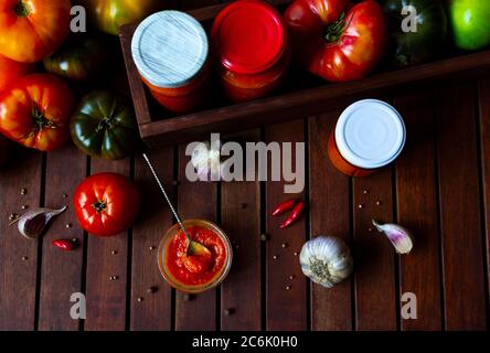 Homemade canned hot tomato sauce adjika Preserve in jars with fresh ingredients - Tomatoes, chilli pepper, garlic, herbs on wooden table. Flat lay Stock Photo