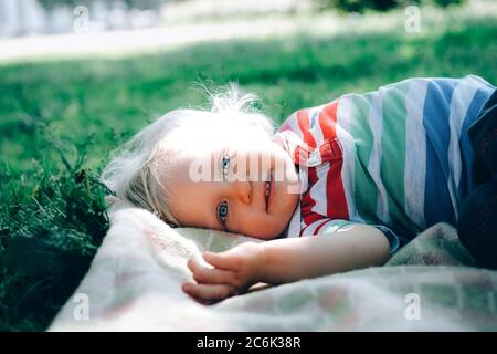 Portrait of a cute happy little boy with blond hair and blue eyes, laying in grass in the park and smiling. Back to normal concept.