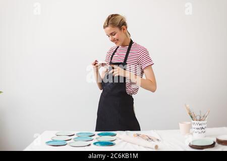 Smiling girl in black apron and striped T-shirt happily taking photos of handmade plates on her cellphone at pottery studio. Stock Photo