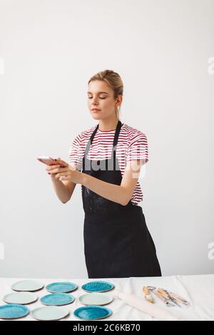 Pretty girl in black apron and striped T-shirt taking photos of handmade plates on her cellphone, spending time at pottery studio Stock Photo