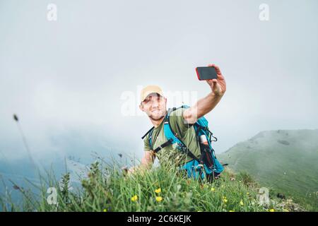 Smiling backpacker man taking selfie picture using smartphone and showing Thumbs Up during walking by the foggy cloudy weather mountain range .  Activ Stock Photo