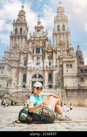 Cheerful smiling Woman backpacker piligrim taking a selfie photo using a modern smartphone siting on the Obradeiro square (plaza) in Santiago de Compo Stock Photo