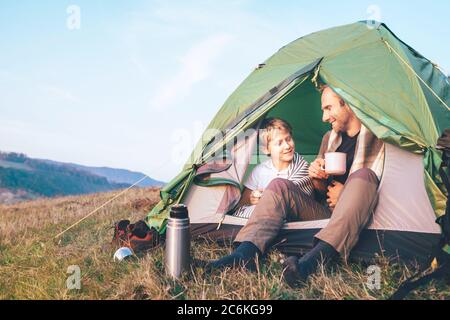 Father and son sit in camp tent drink hot tea and have conversation. Leisure time with father, parenthood Stock Photo