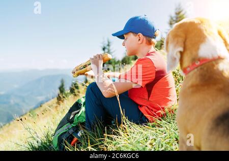 Little smiling boy weared baseball cap enjoying a huge baguette sandwich and his beagle dog friend watching it during a mountain hiking resting snack Stock Photo