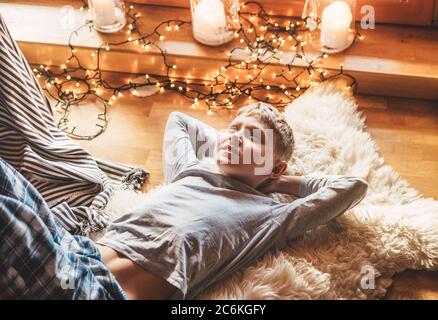 Boy lying on the floor and smiling on sheepskin in a cozy home atmosphere and dreaming about Christmas presents. Peaceful moments of cozy home concept i Stock Photo