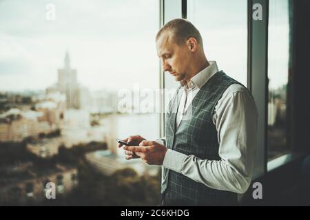 Side view of a serious focused man entrepreneur standing next to the panoramic window of a business office building and using his smartphone Stock Photo