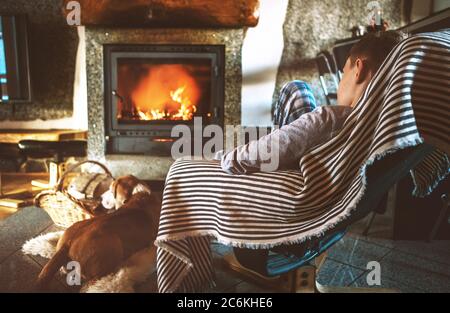 Boy sitting in comfortable armchair in cozy country house near fireplace and enjoying a warm atmosphere and flame moves. His beagle friend dog lying b Stock Photo