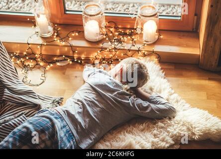 Boy lying on floor on sheepskin and looking in window in cozy home atmosphere. Peaceful moments of cozy home concept image. Stock Photo
