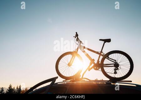Mounted mountain bicycle silhouette on the car roof with evening sun light rays background. Safe sport items transportation using a car concept image. Stock Photo