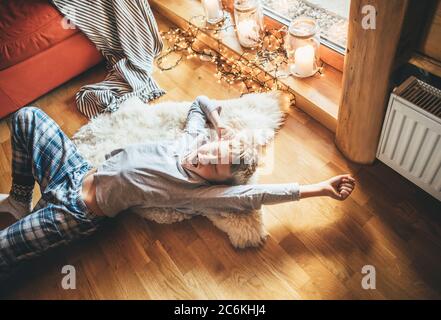 Boy stretching himself lying on floor on sheepskin and looking in window in cozy home atmosphere. Peaceful Lazy moments in cozy home concept image. Stock Photo
