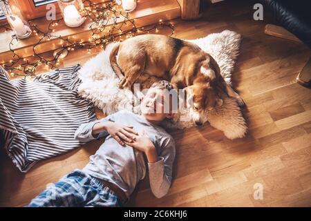 Boy lying on the floor and near beagle dog sleeping on sheepskin in cozy home atmosphere. Peaceful moments of cozy home, holiday time concept image