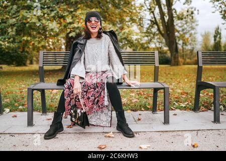 Sincerely laughing female dressed fashion boho style colorful long dress, warm knitted sweater with black leather biker jacket and Beanie Hat with fla Stock Photo