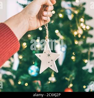 Close-up image of a female hand hanging Christmas wooden star decoration on a spruce tree. Unfocused green branches bokeh with new year lights on the back Stock Photo