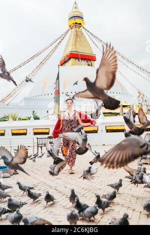 Beautiful female sincerely smiling and going by square through pigeons flock with Boudhanath Stupa - the largest spherical stupas in Nepal. Traveling Stock Photo