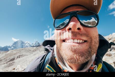 Ultra-wide lens angle portrait shot of high altitude mountain smiling unshaven happy hiker in baseball cap with snow peaks and mountain range beside h Stock Photo