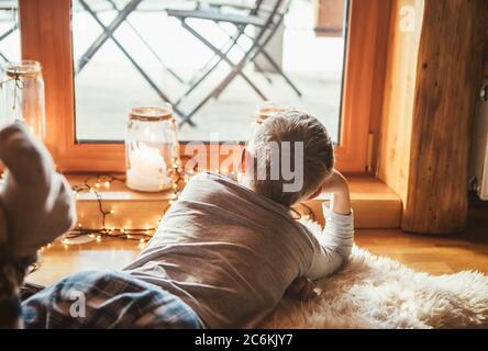 Boy lying on floor on sheepskin and looking in window, lost in dreams or boring. Peaceful moments of cozy home concept image. Stock Photo