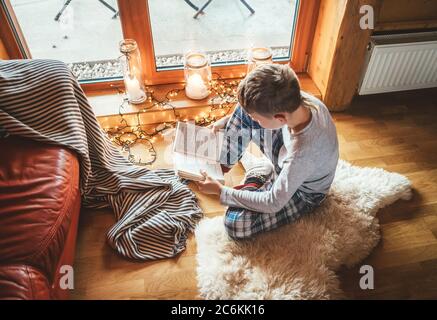 Boy reading book on the floor on sheepskin in cozy home atmosphere. Peaceful moments of cozy home concept image. Stock Photo