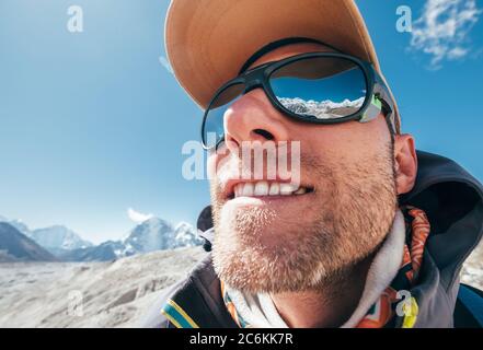 Ultra-wide lens angle portrait shot of high altitude mountain smiling unshaven happy hiker in baseball cap with snow peaks and mountain range beside h Stock Photo
