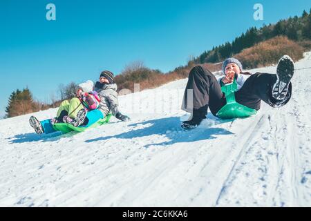 Happy laughing kids sliding down from the snow slope riding sleighs. Funny winter holidays spending concept image. Stock Photo