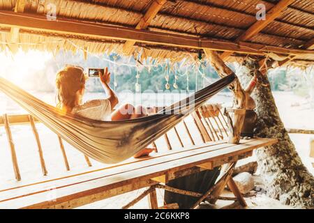 Young female lying in hammock and taking a landscape photo using modern smartphone on Samui island in Thailand. Careless summertime exotic vacation co Stock Photo