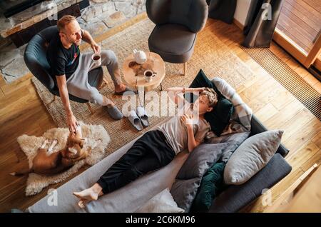 Father and son together at the home living room. Boy lying in comfortable sofa and his daddy stroking their beagle dog and smiling to son. Peaceful fa Stock Photo