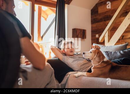 Cozy family tea time. Father and son at the home living room. Boy lying on comfortable sofa and  stroking their beagle dog and smiling. Peaceful famil Stock Photo