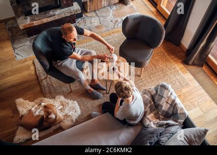 Father and son together at the home living room. Boy sitting on comfortable sofa and his daddy pouring hot tea into white cups. Beagle dog lying on sh Stock Photo
