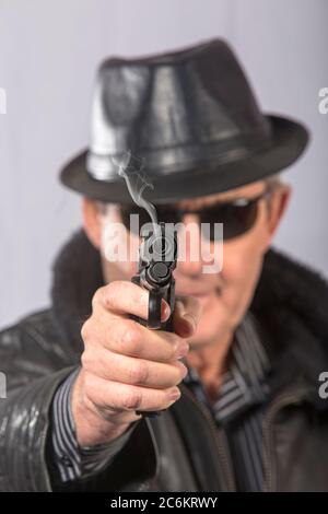 Elderly man with jacket, hat, and black glasses aiming straight ahead with gun from which smoke comes out (Select focus) Stock Photo