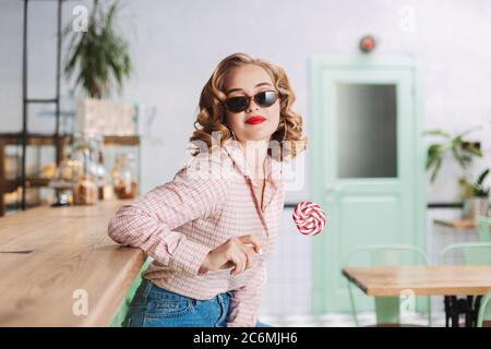 Young pretty lady in sunglasses sitting at the bar counter with lollipop candy while spending time in cafe. Stock Photo